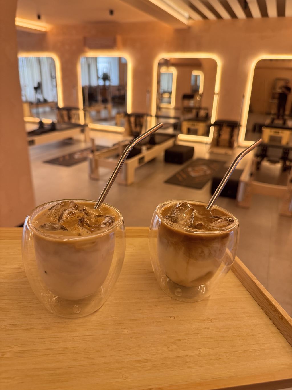 Two iced coffees on a tray with the reformer floor and lit arched mirrors behind